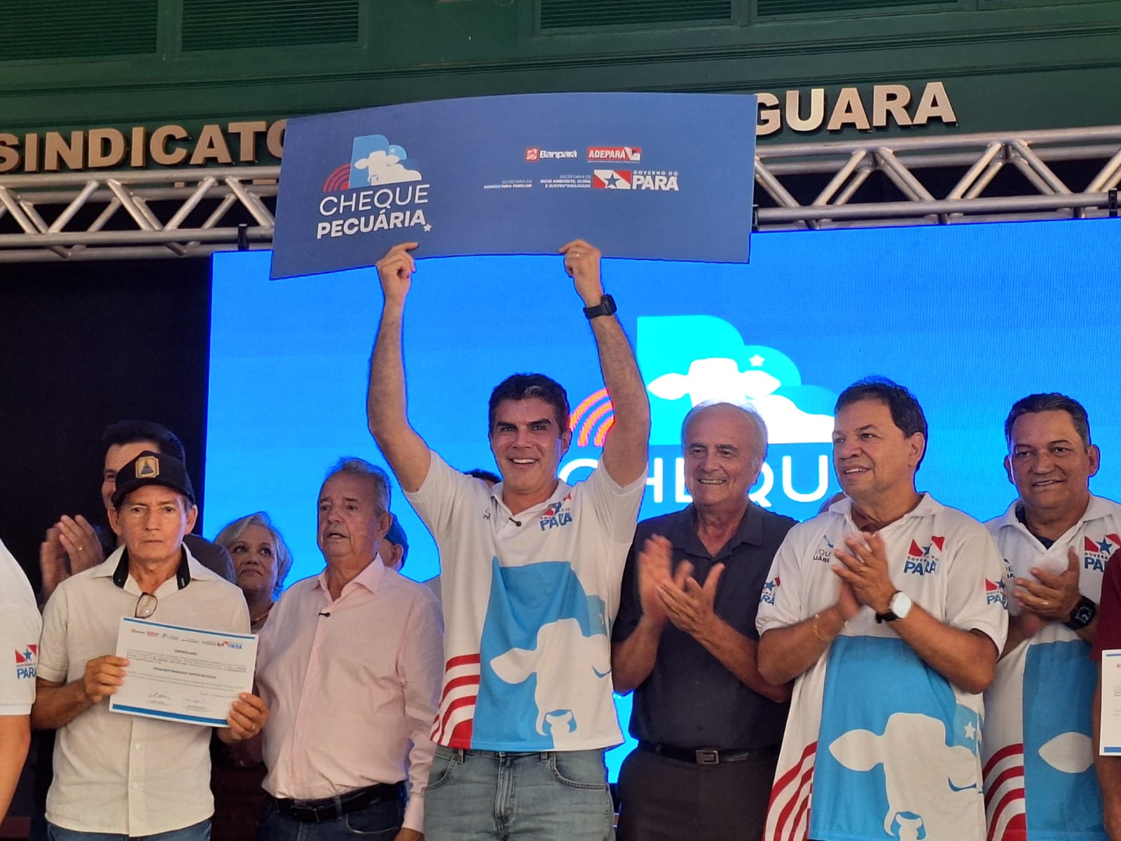 Governador Helder Barbalho exibe o cheque pecuária no alto da cabeça no palco, com pessoas ao lado