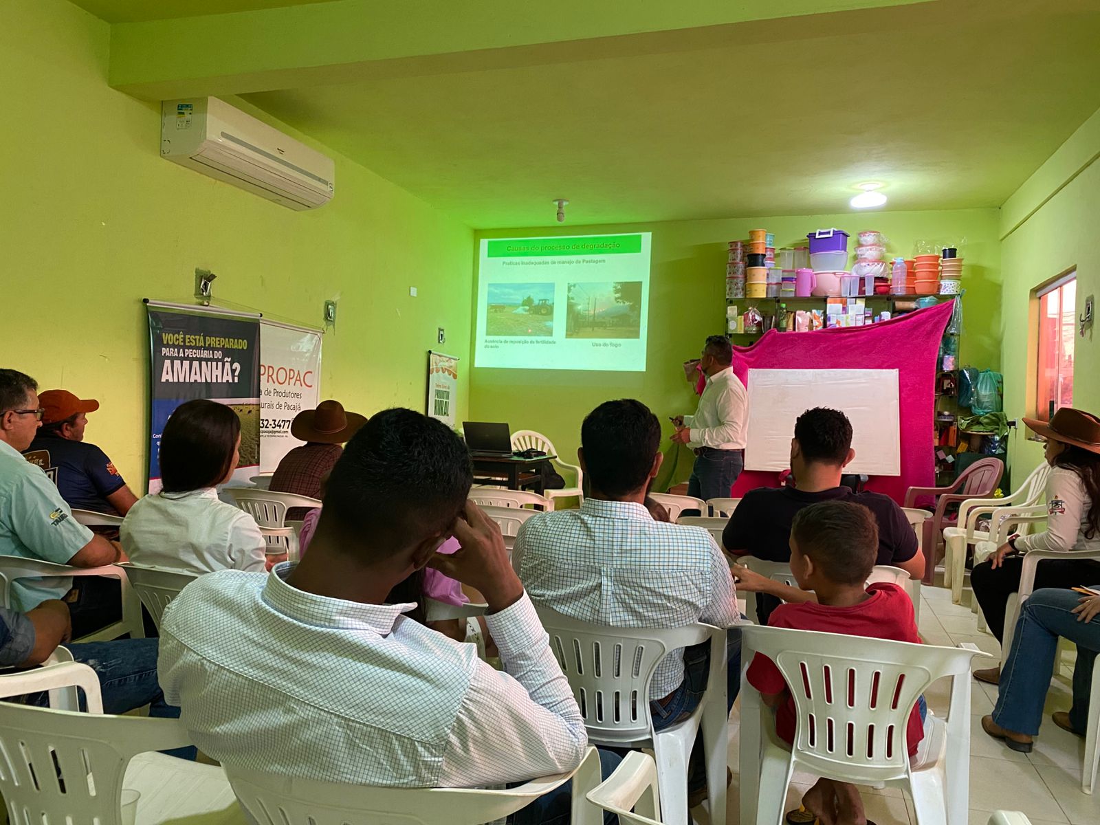 Imagem mostra pessoas de costas sentadas em cadeiras brancas, assistindo curso e professor em pé apontado para projeção de vídeo na parede