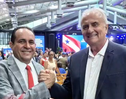 Imagem mostra dois homens brancos, um mais alto e de cabelos brancos, ambos vestidos de paletó dando as mãos em um evento público