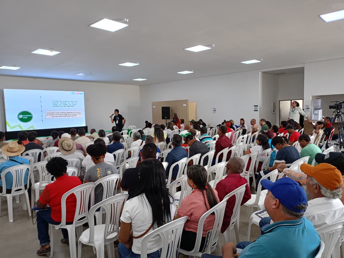 Imagem mostra pessoas sentadas em cadeira assistindo atinvidade com homem ao microfone e telão com informações