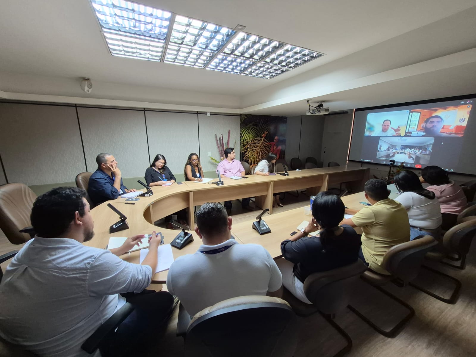 Imagem mostra 11 pessoas em uma mesa circular em reunião olhando para telão com participantes virtuais