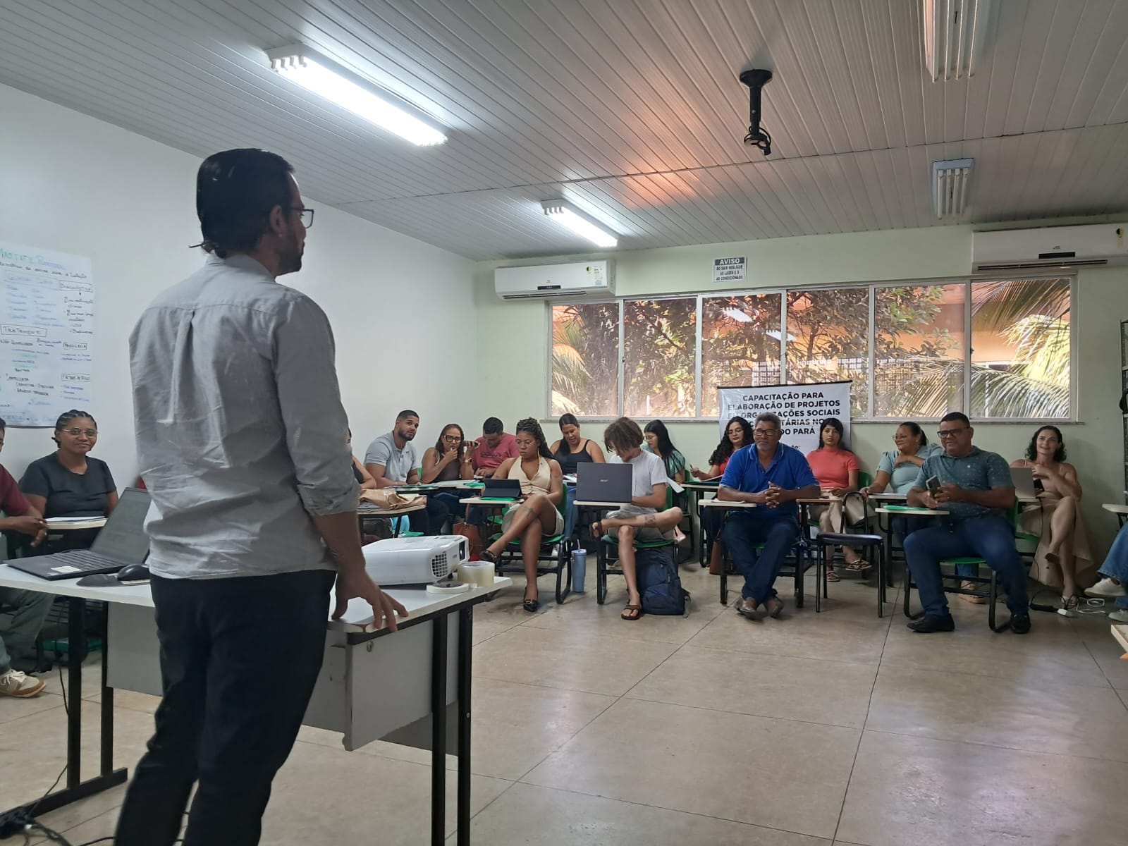 Imagem mostra sala de aula com alunos pessoas sentadas e um homem de pé à frente