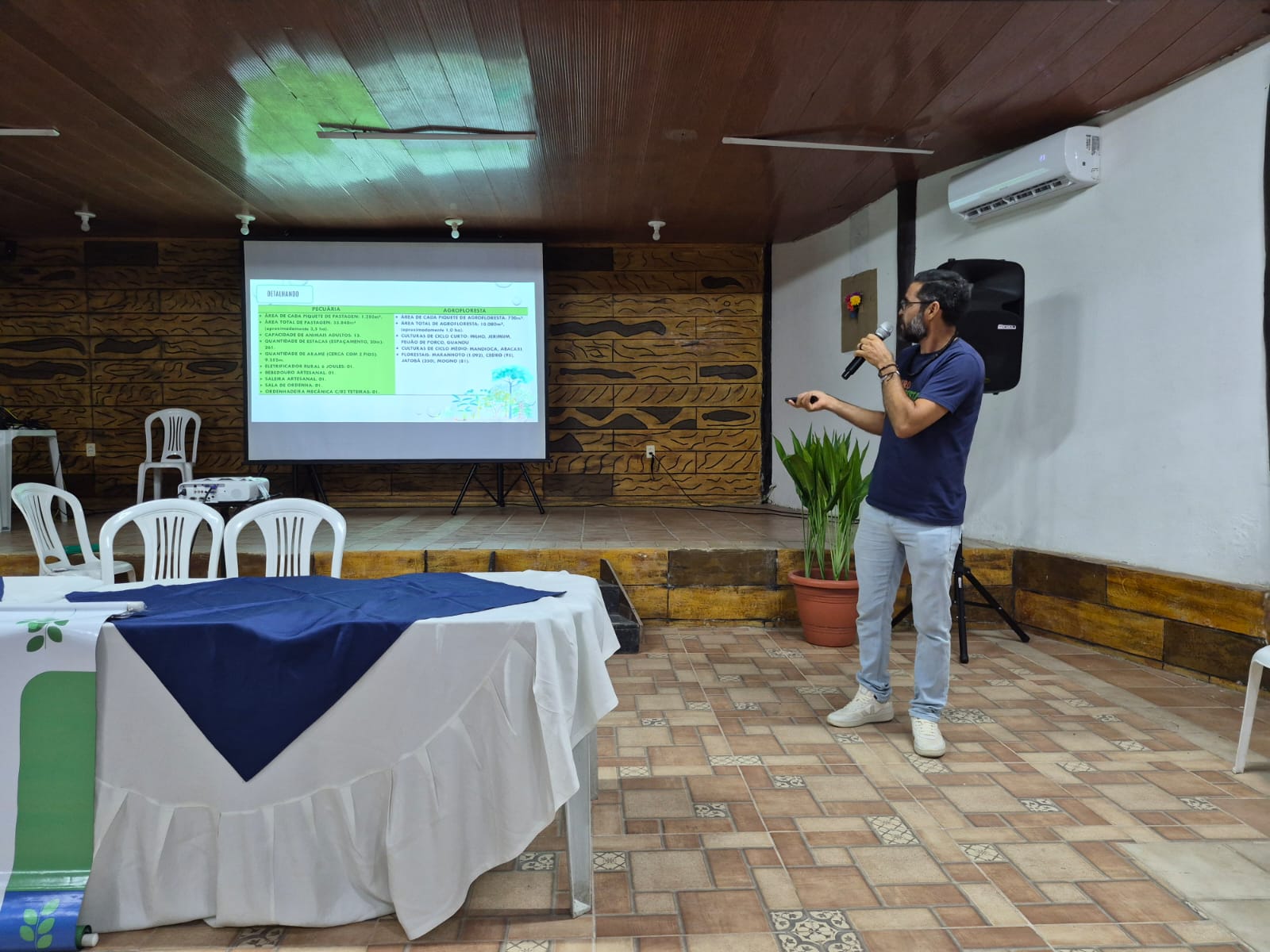 Imagem mostra homem de meia idade, baixo, magro, de camisa azul, ao microfone, apresentando palestra com projeção de imagens atrás dele