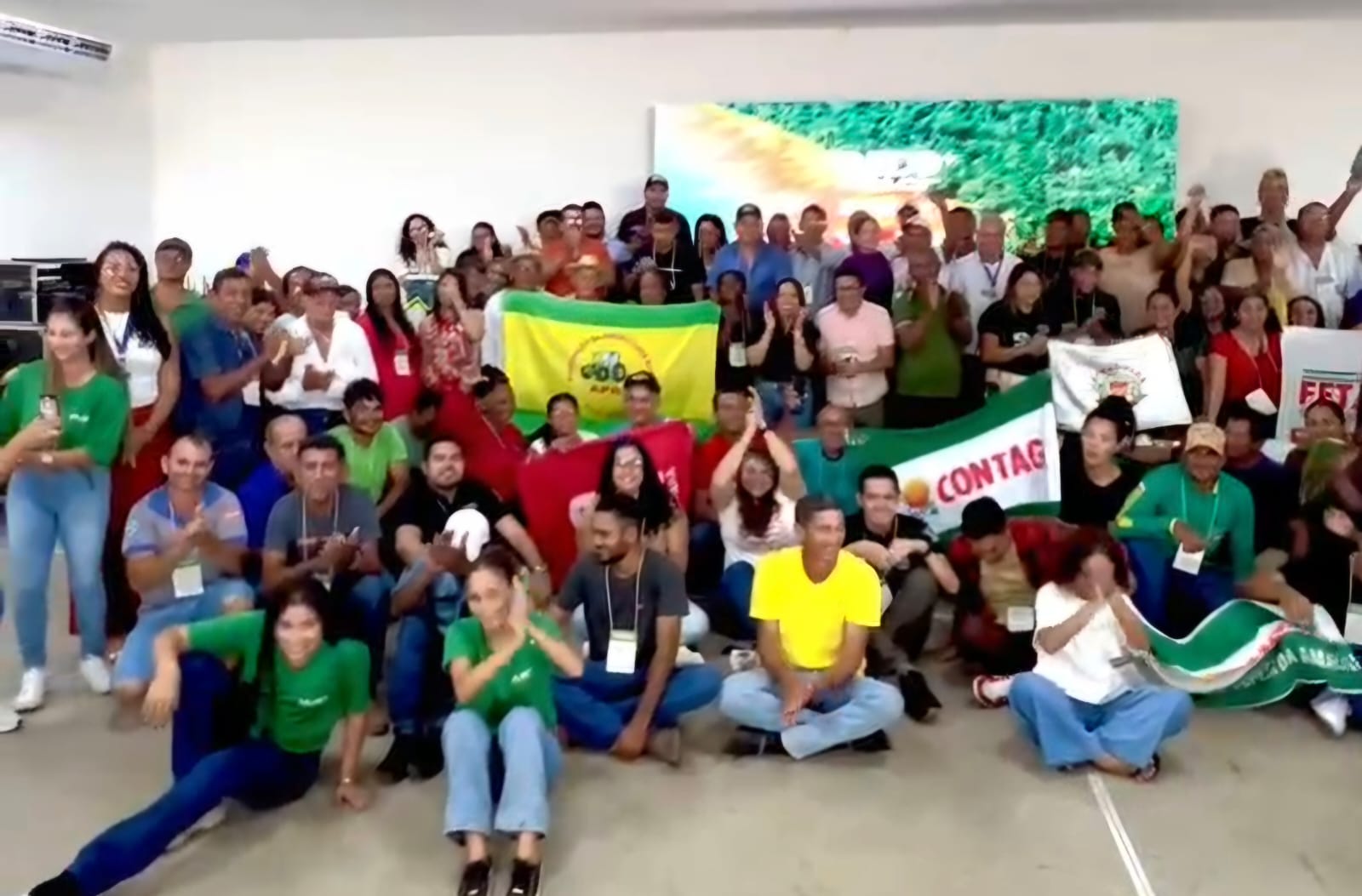 Imagem mostra dezenas de pessoas, algumas de pé, outras sentadas,, algumas com bandeiras na mão, de idades e etinias diversas