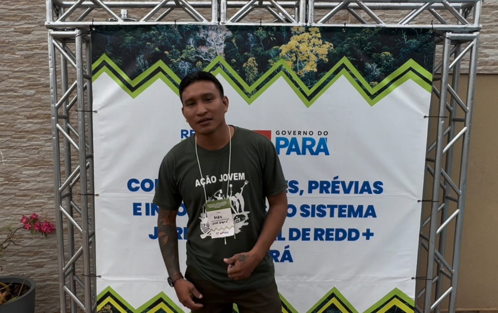 Imagem mostra homem jovem pardo, camisa marrom claro, de pé, gesticulando