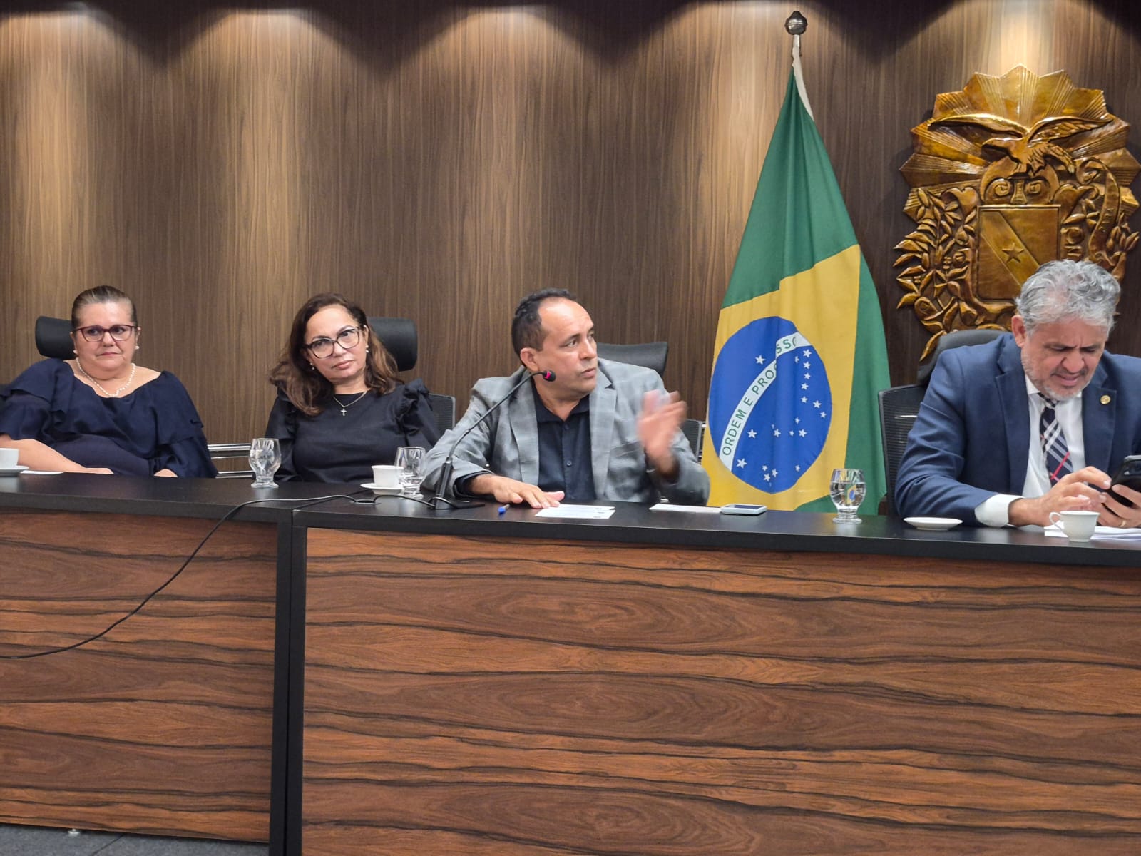 imagem mostra homem ao microfone em bancada ao lado de duas mulheres e um homem