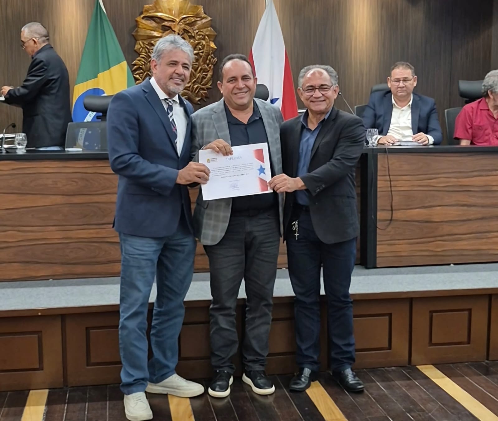 Imagem mostra três homens, o ao centro recebendo um diploma de homenagem, os três sorridentes
